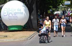 Mulher empurra cadeira de rodas com criança; ao lado, atrás, casal passeia em avenida; à esquerda, balão inflável com os dizeres: "BH é da Gente".