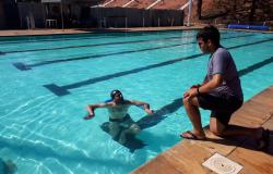 Universitário em piscina com raia olímpica, acompanhado por rapaz fora da piscina, durante o dia. 