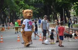 Homem fantasiado de urso com camiseta com os dizeres: "I love BH" passeia em rua com crianças brincando acompanhadas por adulto, durante o dia. 