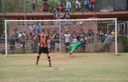 Jogador, de uniforme laranja e preto, dá chute a gol, enquanto goleiro de verde pula no ar para pegar a bola. 