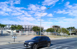 Fachada do Mineirão. Céu azul com algumas nuvens, árvores ao redor do estádio, faixa de pedestre com um carro preto passando no mesmo instante. 