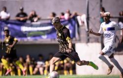 Em um campo de futebol society, um jovem atleta com uniforme preto e amarelo prepara o chute com a perna esquerda, enquanto a bola está à sua frente. Ao lado, um jogador de uniforme branco e azul acompanha a jogada. Ao fundo, desfocados, aparecem outros atletas, integrantes das equipes e parte da arquibancada. A partida acontece durante o dia, sob forte iluminação solar.