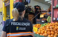 Agente de fiscalização da Prefeitura de Belo Horizonte realiza vistoria em estabelecimento comercial. Ao fundo, são exibidos produtos e frutas à venda.