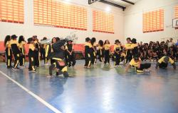 A foto mostra crianças e adolescentes participando de uma apresentação de dança urbana, com movimentos de hip-hop, em uma quadra esportiva coberta. Os participantes vestem uniformes pretos e amarelos, alguns com chapéus, e realizam diferentes movimentos coreográficos, enquanto outros aguardam sua vez. Ao fundo, um grupo de estudantes acompanha a apresentação, sentado e em pé junto à parede.