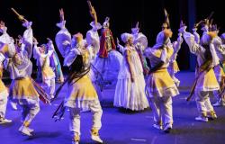 Mulher de vestido e turbante brncos no meio do palco, ao redor dela, homens de roupas brancas e amarelas dançam de maneira coordenada. 