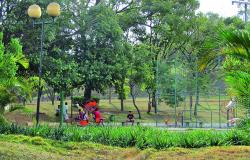 Parque com quadra poliesportiva e aparelhos de ginástica, com pessoas transitando, cercadas de verde, durante o dia. 