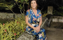 Diretora de Gestão e Educação Ambiental - Laura Maria Moraes Mourão, posa em uma fotografia ao lado de plantas e um vestido azul estampado de plantas.