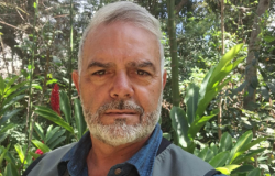 Homem branco, barba e cabelo grisalhos, árvores e plantas ao fundo