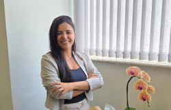 Mulher sorridente, vestindo blazer bege sobre blusa azul, com os braços cruzados, em um ambiente de escritório com persianas ao fundo e uma orquídea sobre a mesa.