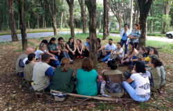 Cerca de vinte pessoas, sentadas no chão, em círculo, ao redor de uma árvore, em região arborizada, durante o dia. 