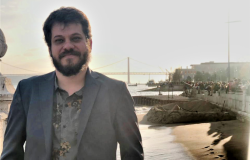 Homem de blazer sorri; no fundo, uma praia e uma ponte, durante o dia. 