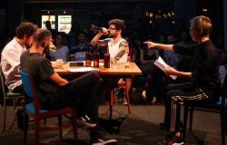 Quatro homens conversam em uma mesa de bar cênica, em um palco