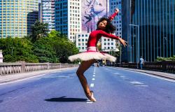 Mulher negra com blusa rosa e saia branca de bailarina faz pose de ponta no viaduto Santa Tereza, durante o dia. 
