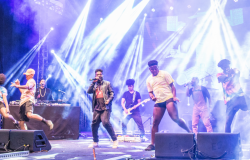 Grupo afro com DJ, guitarrista, vocalista e quatro dançarinos se apresenta em palco. 