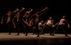 Seis homens e uma mulher, agrupados em um grupo de quatro e um de três, dançam à meia luz, em palco. 