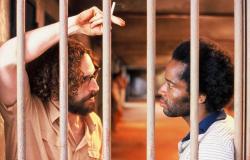 Homem branco, barbudo e com cigarro na mão olha para homem negro; ambos estão atrás das grades. Cena do filme "Quase dois irmãos".