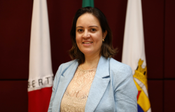 Mulher sorrindo, com blusa azul clara posa em frente as bandeiras de BH e de MG.