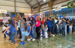 #paratodosverem: a imagem mostra um grupo animado de crianças e adultos participando de uma gincana em uma quadra esportiva. Alguns(mas) levantam os braços e seguram pompons e fitas azuis. O ambiente está decorado com faixas e a bandeira do Brasil. Ao fundo, uma faixa diz "Descobrindo a nossa mineiridade com a Emei Mariquinhas – 2025".