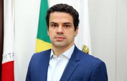 Castellar Modesto Guimarães Neto (EXONERADO) Homem de terno azul e camisa branca posa em frente às bandeiras de Minas, do Brasil e da PBH
