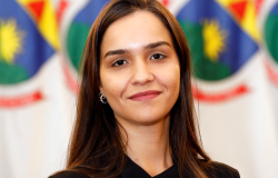 Mulher de pele clara, com cabelos longos, lisos e castanhos repartidos ao meio. Tem sobrancelhas marcadas, usa pequenos brincos prateados e veste blusa preta sem mangas. Está de frente para a câmera, com um leve sorriso e expressão serena.