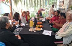  A imagem, em ambiente externo, mostra um grupo de cerca de dez mulheres reunidas ao redor de uma mesa, participando de reunião acompanhada de um lanche com bolo, pães, frutas e garrafas térmicas.