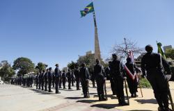 Guardas municipais de Belo Horizonte vestem uniforme azul marinho e estão alinhados em formação durante uma cerimônia ao ar livre na praça da Bandeira. No centro, uma bandeira do Brasil tremula no alto de uma haste sob o céu limpo. 