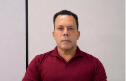 Homem, sem barba, usando uma camisa polo vermelha vinho, sentado em uma cadeira de escritório em uma sala branca