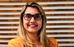 Mulher sorridente, pele clara, cabelo loiro e liso, usa óculos e camisa amarela.