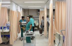 TRês profissionais de saúde, com carrinhos e material hospitalar, carregam equipamentos em meio a macas de hospital, em sala do Hospital Metropolitano Odilon Behrens. 