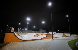 Visão noturna de uma pista de skate ao ar livre, vazia, com rampas de concreto cinza e detalhes laranjas. Vários postes altos iluminam fortemente o local