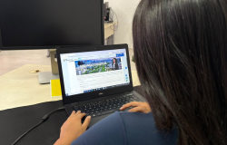 Mulher de cabelos escuros, vista de costas, utiliza um notebook em um ambiente de trabalho. Na tela, está aberta uma página do site da Prefeitura de Belo Horizonte com informações sobre mudanças climáticas.