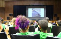  Plateia atenta de estudantes reunida em um auditório acompanhando uma apresentação na tela principal. Em primeiro plano, destaca-se a diversidade do grupo com o visual vibrante de um cabelo cacheado com tons de roxo e rosa.