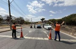 Agentes da Prefeitura de Belo Horizonte trajando de colete laranjado, ao lado de cones, sinalizando com braços erguidos para veículos parados em via de trânsito rápido