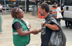 A foto mostra uma cena ao ar livre, em uma calçada movimentada de uma área urbana. No primeiro plano, duas pessoas conversam e apertam as mãos. À esquerda, uma mulher usa colete verde com faixas refletivas e segura uma garrafa de água. À direita, um homem veste camiseta preta e parece estar usando uma capa ou plástico escuro sobre os ombros; ele também segura uma garrafa de água. Ao fundo, há ônibus, veículos, agentes uniformizados, grades de isolamento e equipamentos de sinalização.