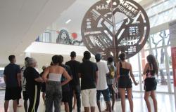 Em ambiente interno amplo e iluminado, um grupo de jovens e adultos, vistos de costas, observa uma grande escultura metálica em formato de árvore. A obra ocupa o centro da imagem e traz palavras e expressões recortadas em sua estrutura, entre elas “mãe”, “memória” e “juventude negra viva”. Ao fundo, há mezanino, paredes claras e grandes janelas de vidro, que permitem a entrada de luz natural.