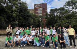 #paratodosverem: na imagem, grupo de estudantes e professores(as) posa para foto em área externa, com vegetação ao fundo e um prédio com a inscrição “bh.tec” ao centro da imagem. Os(As) estudantes demonstram animação, com gestos, acenos e sorrisos para a câmera. O céu está parcialmente nublado.