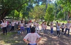 Um grupo de pessoas realiza uma prática corporal Lian Gong ao ar livre. Todos estão em pé com as mãos apoiadas na cintura, mantendo a mesma postura de exercício.