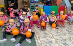 #paratodosverem: na foto, crianças pequenas estão sentadas em um chão com piso de cerâmica marmorizada, usando mochilas e coroas de papel na cabeça. Elas seguram bexigas laranja e vestem roupas de frio coloridas. Ao fundo, duas educadoras acompanham o grupo em ambiente com sofá, casinhas de madeira e outros brinquedos.