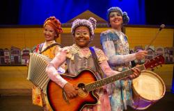 Três atrizes do espetáculo "Mamulenga", do Grupo Girino, sorriem no palco segurando instrumentos musicais. A da esquerda toca acordeão, a do centro toca violão e a da direita carrega uma zabumba. Elas usam figurinos coloridos, maquiagem artística e cabelos feitos de lã estilizados. 
