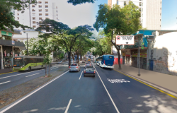 Fotografia de uma via urbana com três faixas de tráfego, árvores no canteiro central e prédios ao fundo. Na faixa da direita, há a pintura no asfalto indicando “ÔNIBUS” e uma placa aérea com os dizeres “Faixa exclusiva para ônibus – 2ª a 6ª, 6h às 9h e 16h às 20h”. Alguns veículos circulam na via, incluindo ônibus e carros.
