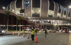 Foto noturna da Arena MRV, estádio do Atlético Mineiro, com a fachada iluminada e o escudo do clube em destaque. Na rua em frente, agentes de trânsito organizam a circulação de veículos com o auxílio de cones.