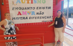 #paratodosverem: fotografia de duas mulheres sorrindo ao lado de um mural em parede vermelha. A frase no mural diz: "AUTISMO — Enquanto existir AMOR, não haverá diferenças!" As palavras "AUTISMO” e "AMOR" estão escritas com letras de cores variadas, e a letra "O" é representada por um coração com estampa de peças de quebra-cabeça, símbolo do autismo. O restante da frase está em preto.