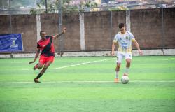 Dois jogadores disputam a bola em uma partida de futebol society, realizada em campo de grama sintética. Um atleta veste uniforme vermelho e preto, enquanto o outro usa uniforme branco e amarelo.