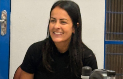 Mulher de cabelos longos e escuros, sorrindo, sentada em uma mesa. Ela veste camiseta preta e está em um ambiente interno, com porta ao fundo e garrafas sobre a mesa.