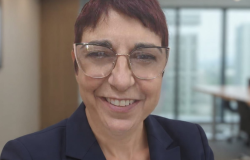 Mulher branca, cabelos curtos e pintados de vermelho, usando blazer azul marinho . O fundo é um escritório comum.