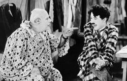 Cena de filme em preto e branco com Charlie Chaplin. A esquerda um personagem sentado com roupas de palhaço, sem peruca e apontando o dedo para a direita em direção ao personagem de Chaplin, que veste uma roupa xadrez e está sentado com as pernas cruzadas. 