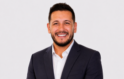 Homem de cabelos curtos e escuros, barba bem aparada, pele clara, olhos escuros e sorriso amplo. Veste camisa branca e blazer escuro, com aparência profissional e descontraída.