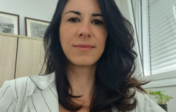 Mulher branca, cabelos e sobrancelhas pretas. Vestindo um blazer branco com riscas de giz preta. Ao fundo um escritório corporativo comum.
