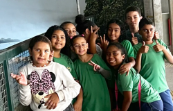 #paratodosverem: a foto mostra um grupo de estudantes em um ambiente interno. A maioria está vestida com o uniforme escolar, composto por camiseta verde e bermuda azul. À esquerda, uma aluna veste blusa branca com estampa da Minnie Mouse e calça preta. O local tem piso de granito e uma divisória metálica à esquerda. 