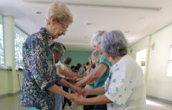 Duas idosas dançam de mãos dadas, olhando uma para a outra, no fundo, mais de oito pares de idosos repetem o gesto. 